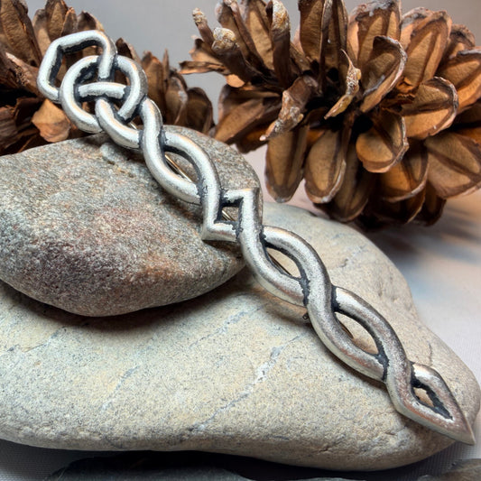 A silver-toned pewter kilt pin with a repeating interlocking link pattern draped over stacked smooth stones, with pinecones in the background on a light neutral surface.