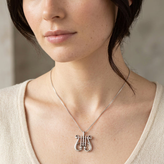 A silver-toned necklace with a lyre-shaped pendant resting on a gray stone slab with natural rocks in the background.