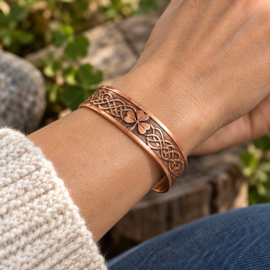 A copper-colored engraved cuff bracelet with Celtic knot patterns and a clover motif, displayed on smooth gray stones with pinecones in the background.