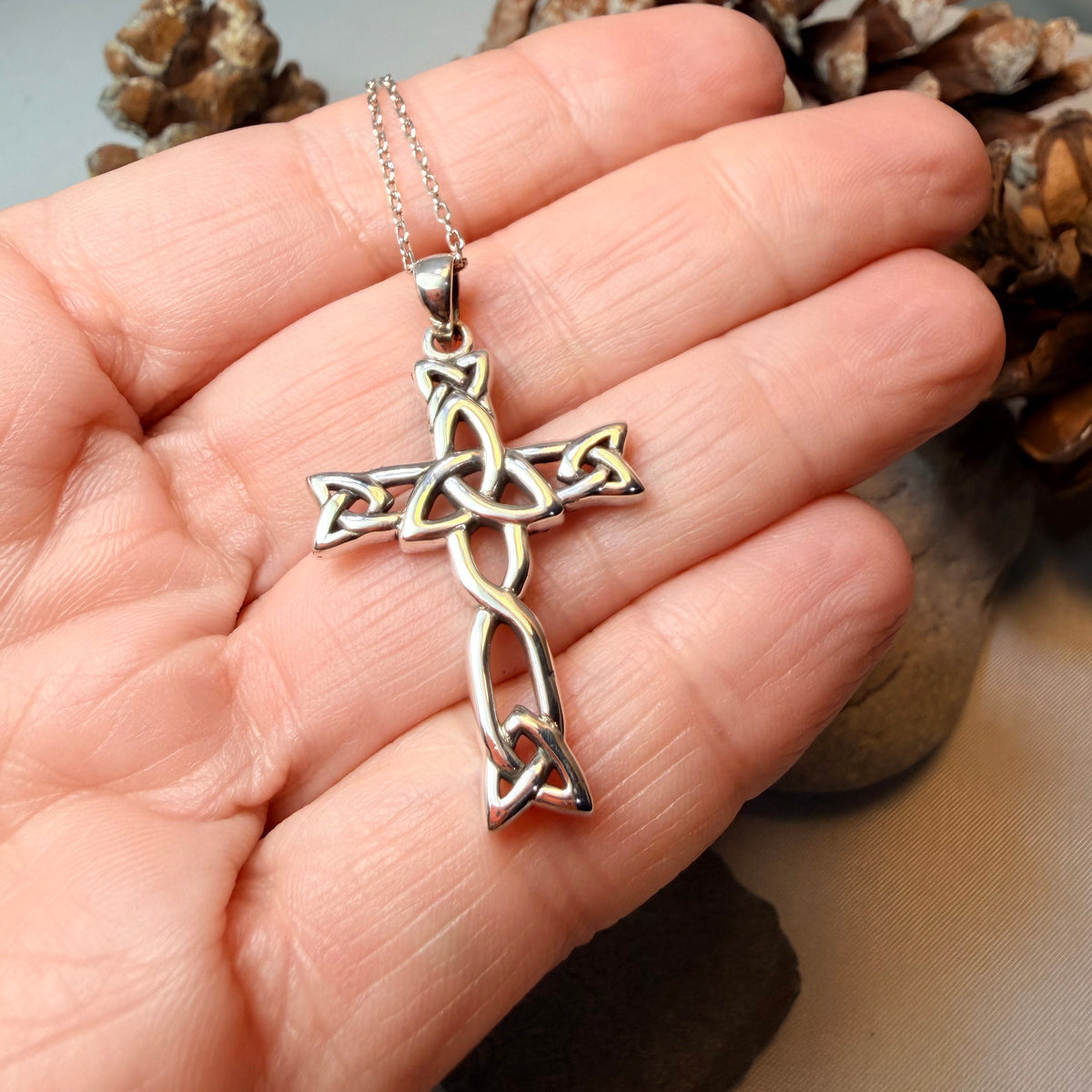A sterling silver Celtic cross pendant on a delicate chain, held in the palm of a hand with pinecones in the background.