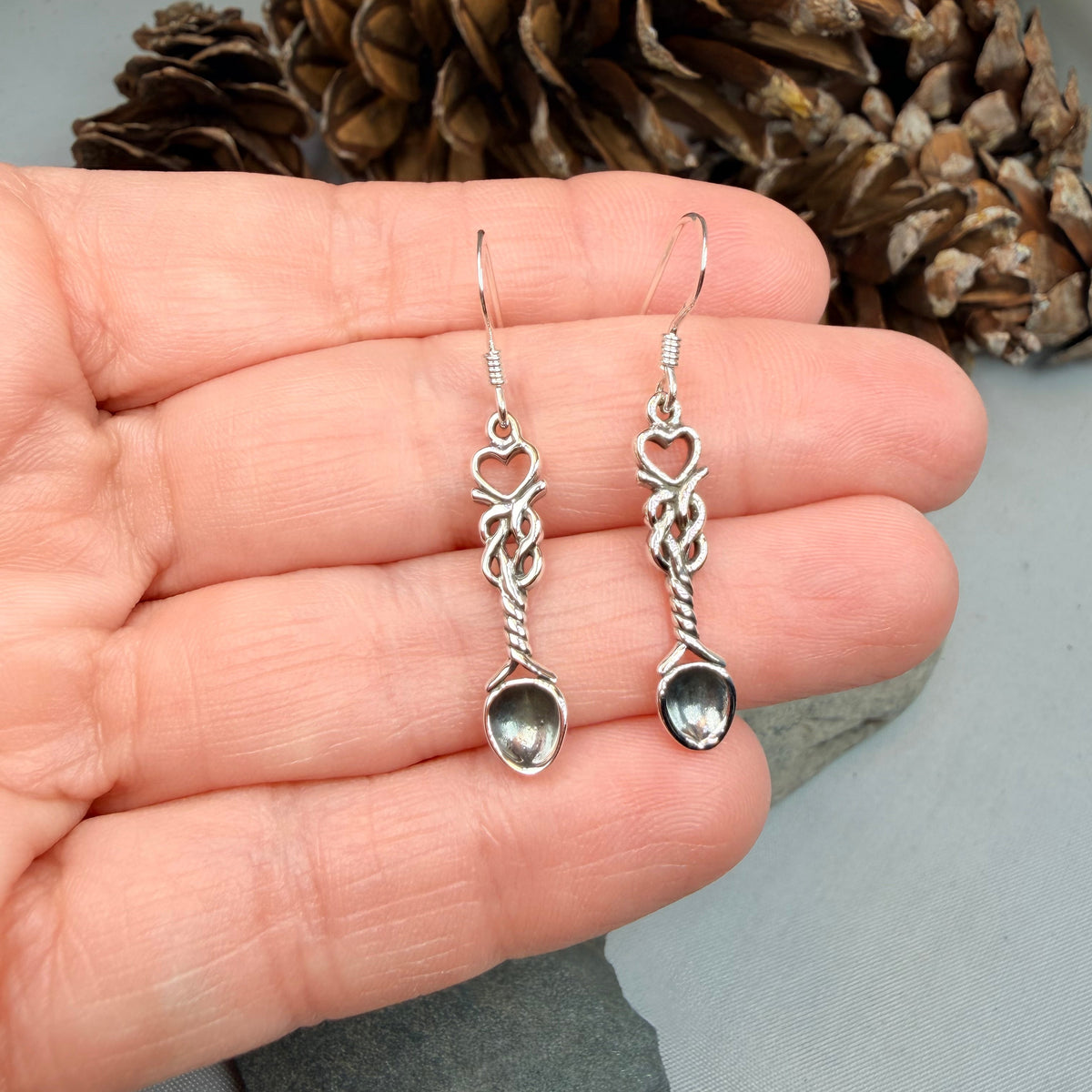 A pair of handmade silver dangle earrings held in a person's hand, featuring knotted wire details and small teardrop-shaped silver pendants, with a pinecone and gray stones in the background.
