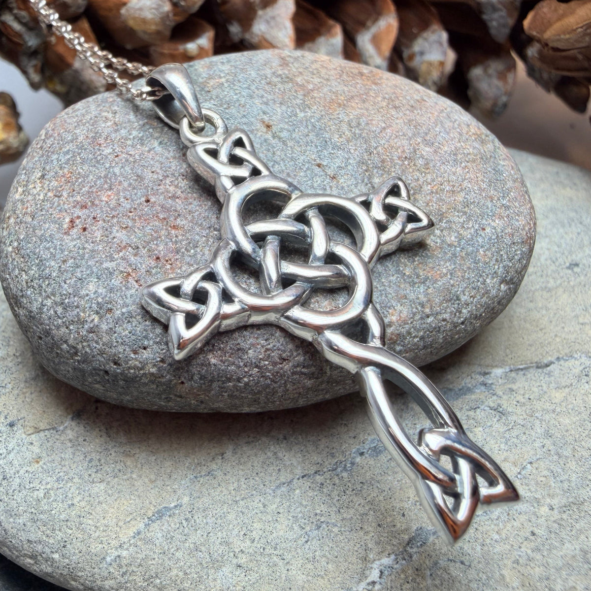 A silver Celtic knot cross pendant with a center dara knot on a matching chain, displayed on smooth gray stones with pinecones in the background.