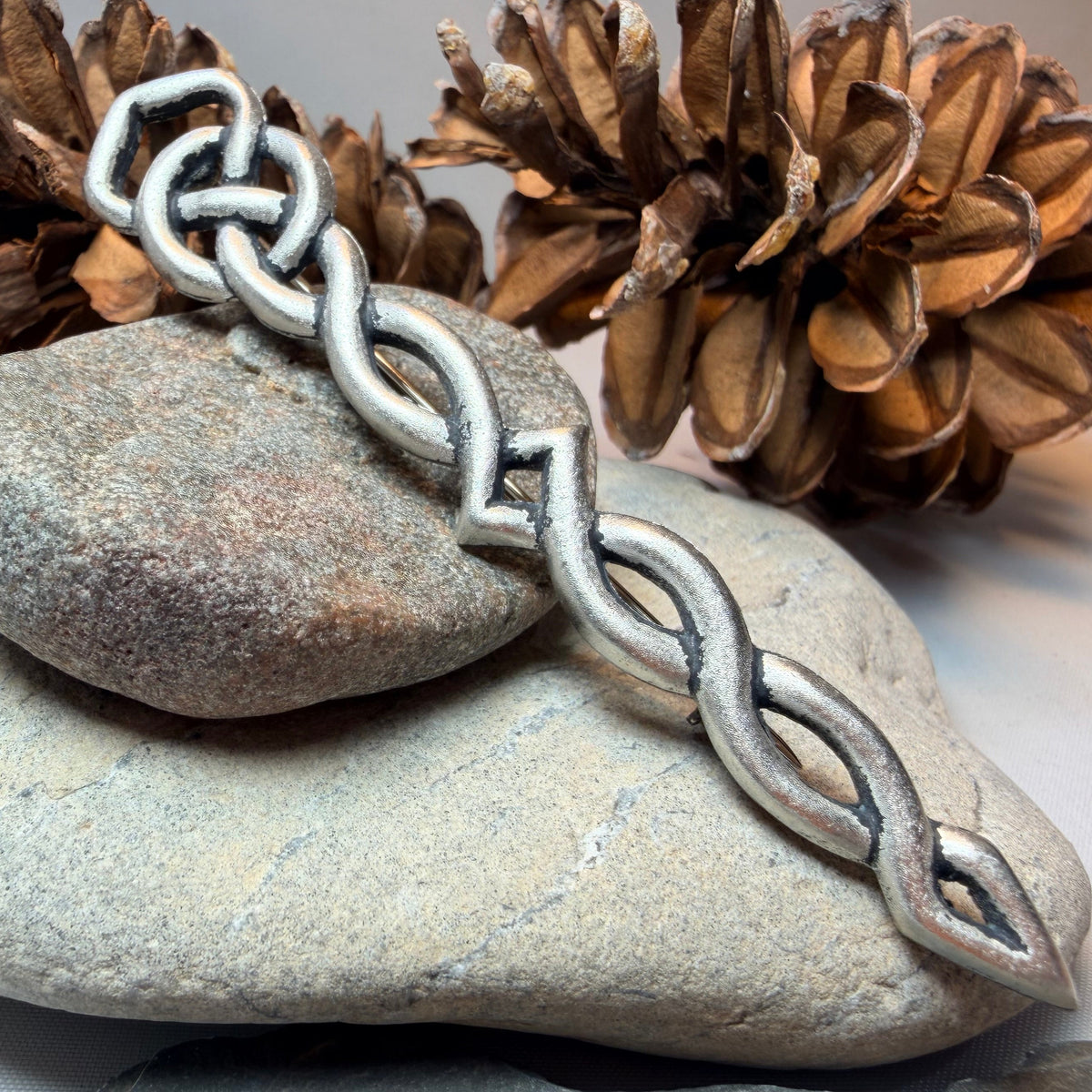 A silver-toned pewter kilt pin with a repeating interlocking link pattern draped over stacked smooth stones, with pinecones in the background on a light neutral surface.