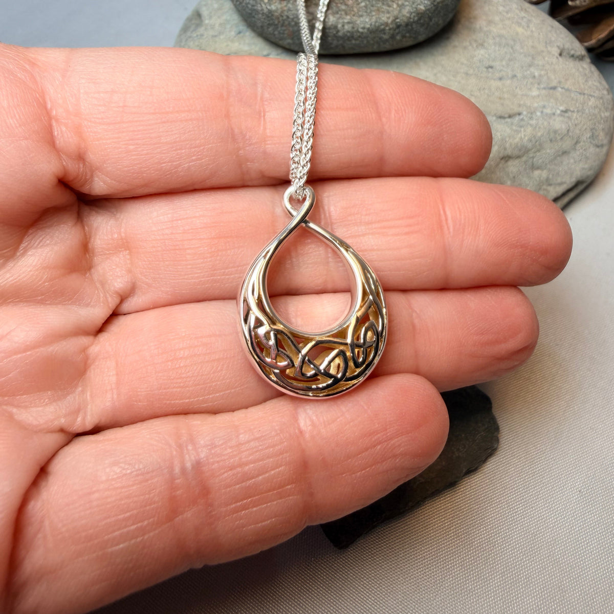 A silver teardrop-shaped pendant necklace with intricate Celtic knotwork detailing, held in a person's hand against a neutral gray background with decorative stones.