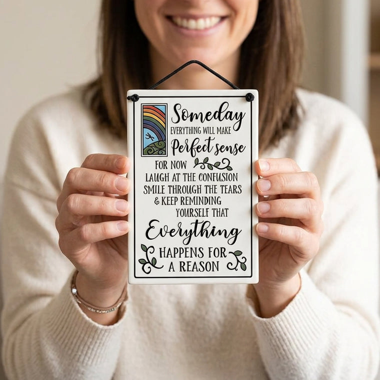 A rectangular white hanging wall sign with a black cord at the top, featuring a humorous quote in black script and serif text: 'Someday everything will make perfect sense for now laugh at the confusion smile through the tears & keep reminding yourself that everything happens for a reason.' A small rainbow illustration appears on the left side.