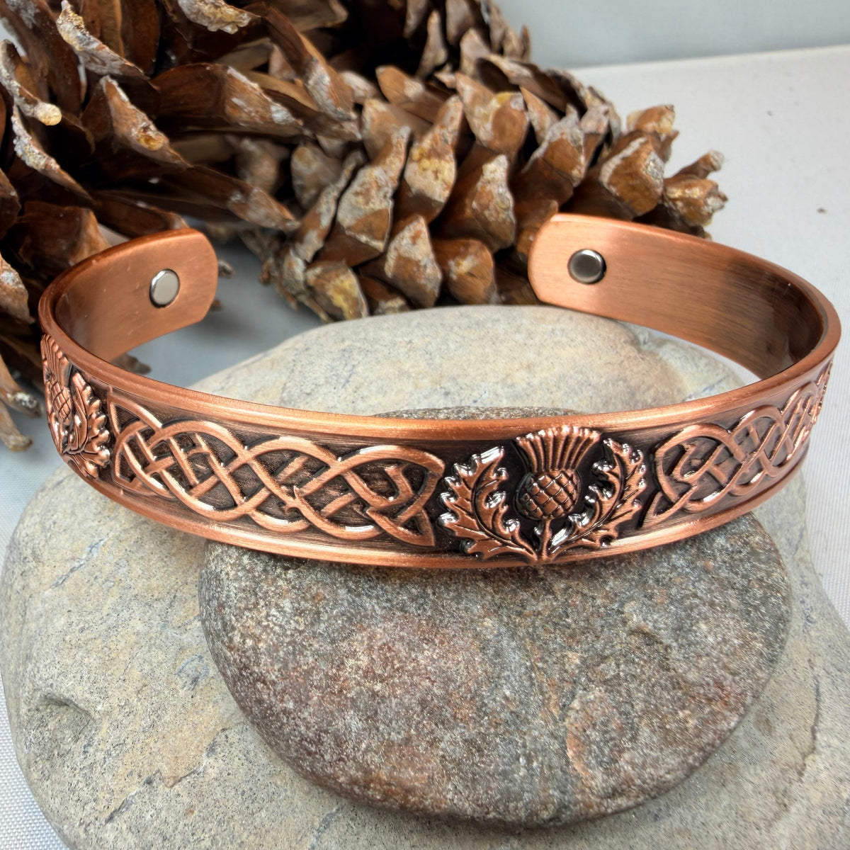 A copper-colored metal cuff bracelet with Celtic knotwork and a thistle emblem, displayed on stacked stones with pinecones in the background.
