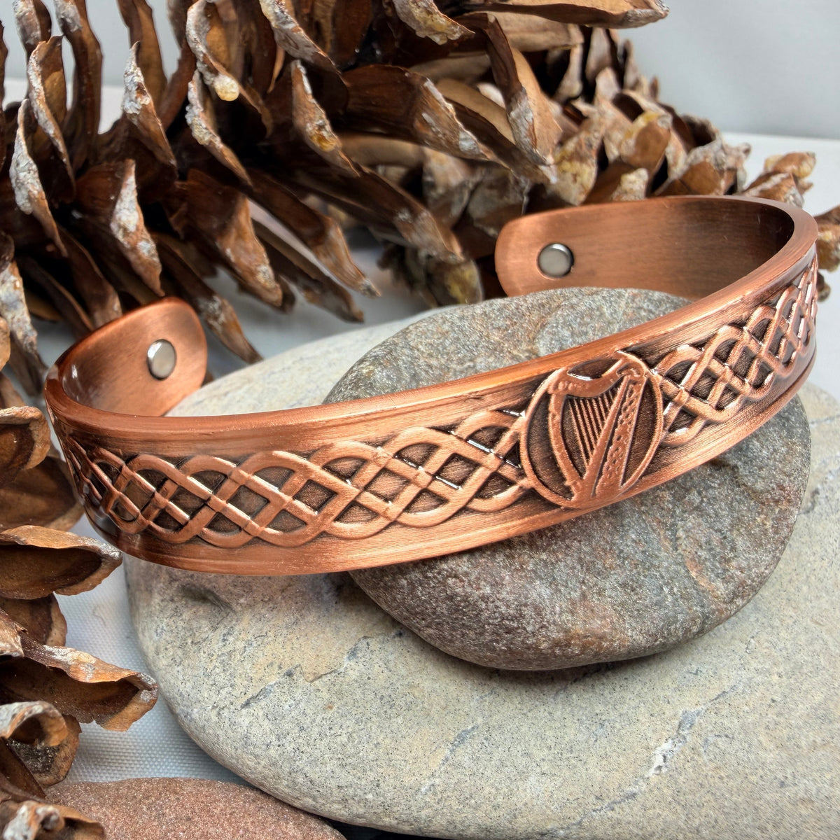 A copper-colored metal cuff bracelet with Celtic knotwork and a harp design, displayed on a stone with pinecones.