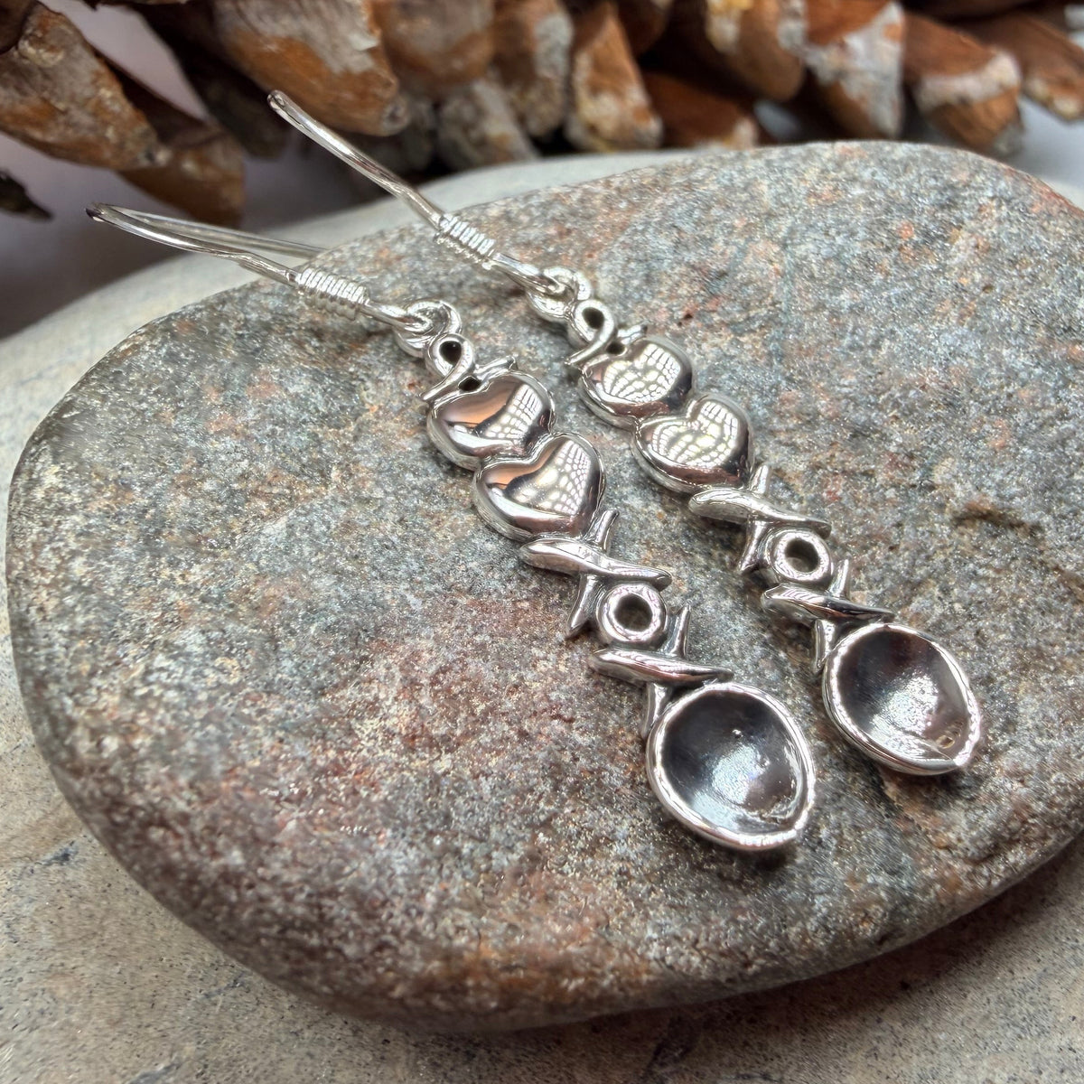 A pair of silver dangle welsh love spoon earrings displayed on a smooth gray stone, with pinecones in the background.