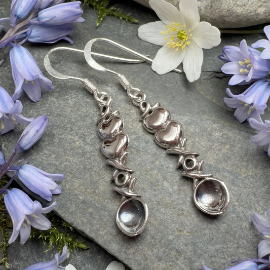 A pair of handmade sterling silver dangle earrings with organic, wavy links and a teardrop-shaped clear stone at the bottom, displayed on a textured gray slate surface.