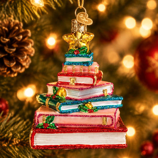 A Christmas ornament shaped like a stack of books, with each book in a different festive color including red, pink, teal, and white. The stack is decorated with glitter, gold trim, small gold stars, holly leaves, and red berries, and topped with a gold star and gold hanging loop, shown on a plain white background.