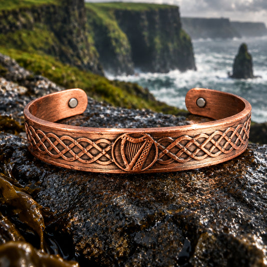 A copper-colored metal cuff bracelet with a Celtic knot pattern and a harp in the center, displayed on a stone with pinecones in the background.