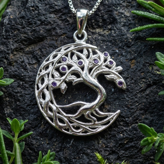 A silver crescent moon and tree of life pendant necklace with small purple gemstone eyes, resting on a smooth gray stone with pinecones in the background.