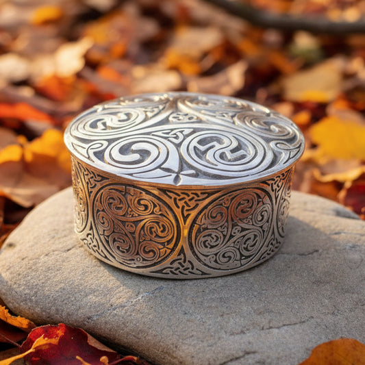 An ornate round silver-toned metal box with intricate Celtic knotwork engravings on the lid and sides, displayed on a stone slab with pinecones in the background.