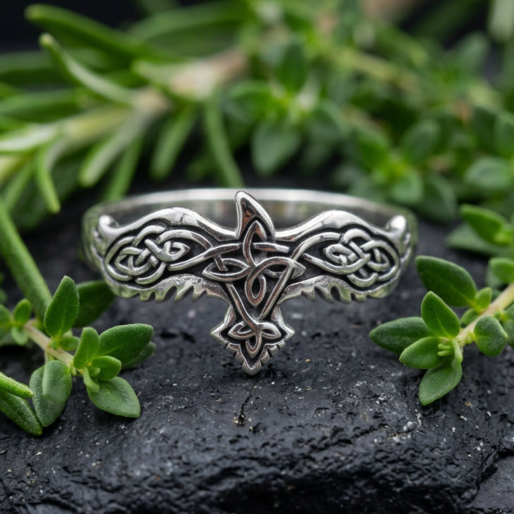 A silver-toned Celtic knot ring with a small rose gold accent, displayed on a gray stone among smooth river rocks.