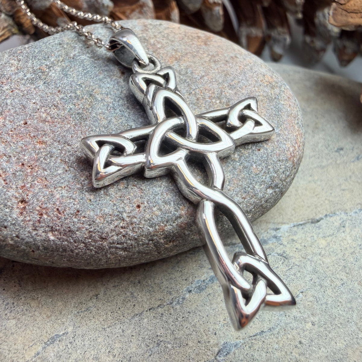 A silver Celtic knot cross pendant on a matching chain, displayed on a smooth gray stone with pinecones in the background.