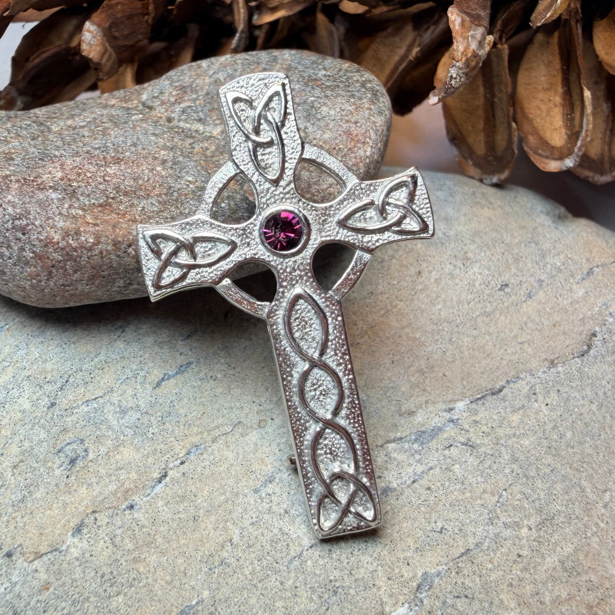 A silver Celtic cross pin with a small purple gemstone at the center, resting on a gray stone with pinecones in the background.