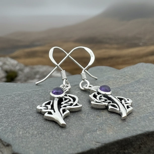 A pair of sterling silver dangle earrings with purple amethyst thistle charms, displayed on a textured gray slate surface.