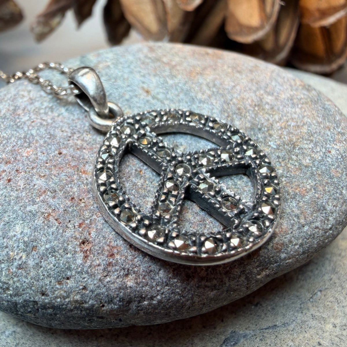 A silver peace sign pendant necklace studded with small clear stones, resting on a smooth gray rock with pinecones in the background.