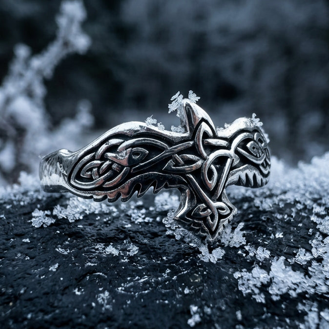 A silver-toned Celtic knot ring with a small rose gold accent, displayed on a gray stone among smooth river rocks.