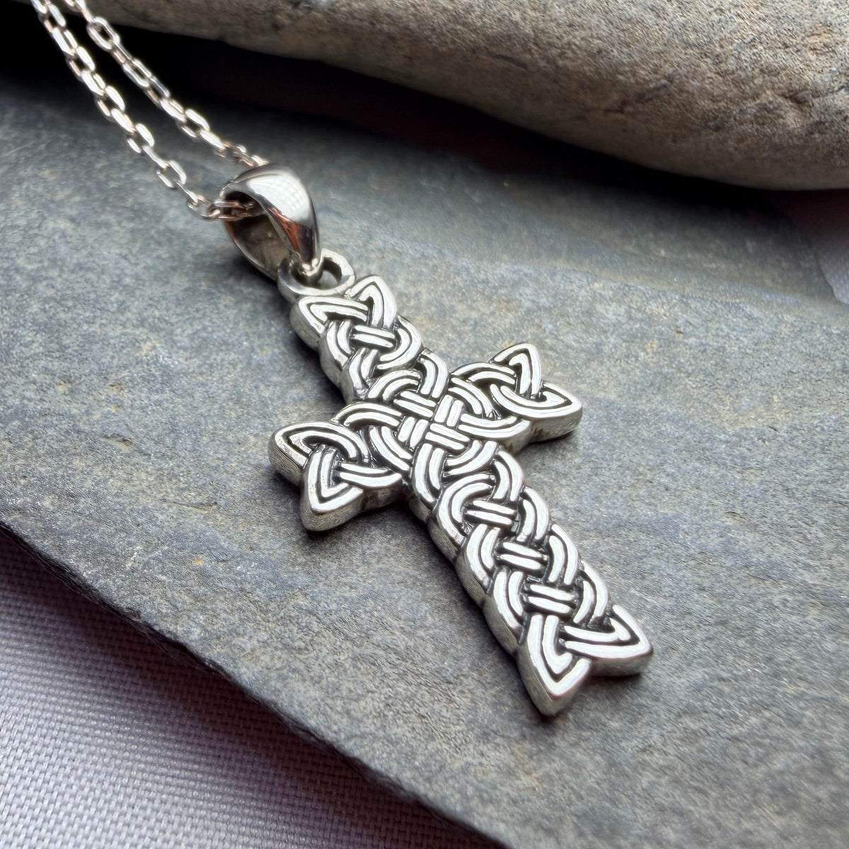 A silver Celtic knot cross pendant necklace resting on a dark gray slate stone, with a smooth beige stone in the background.