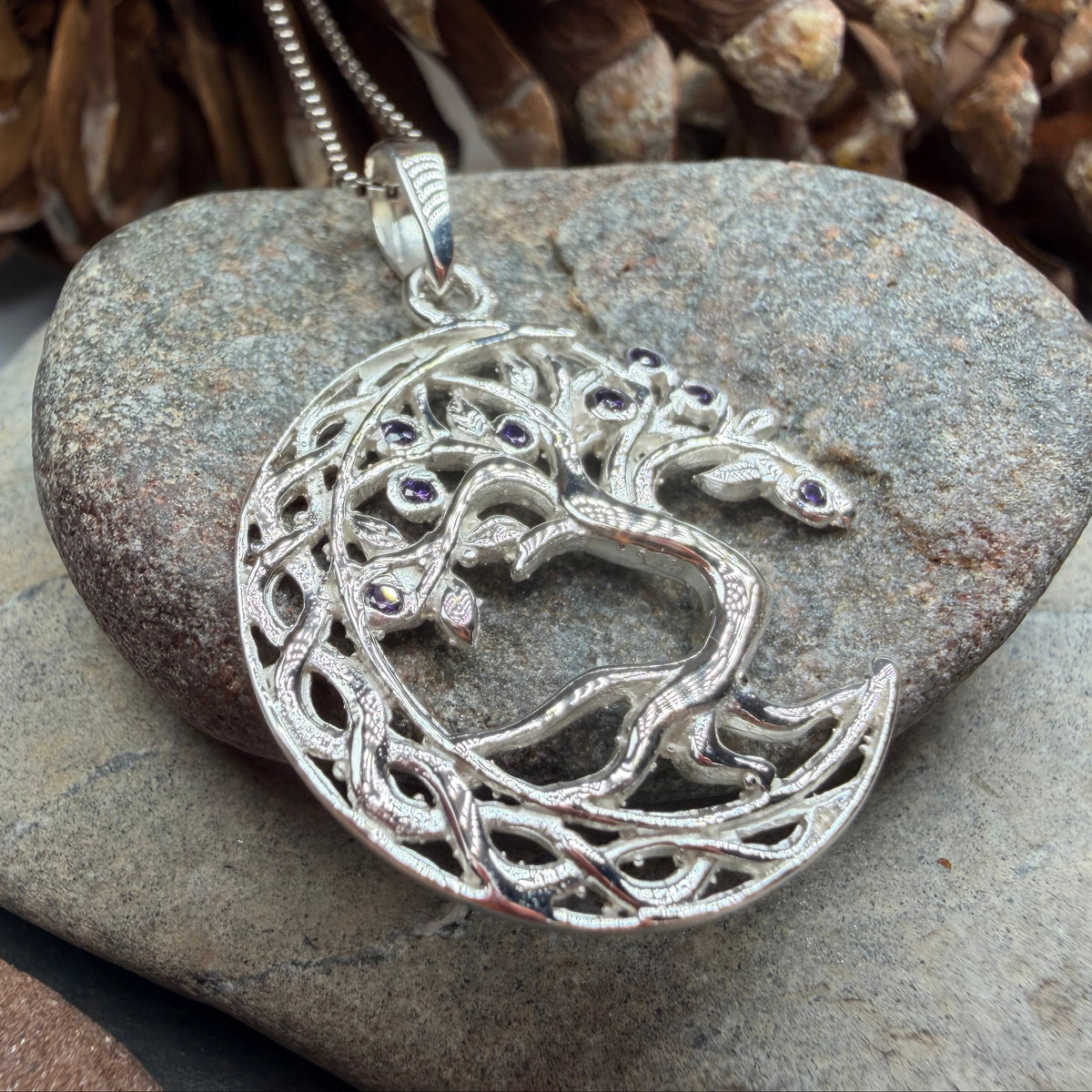 A silver crescent moon pendant necklace featuring a tree of life design with small dark gemstone eyes, resting on a gray stone with pinecones in the background.