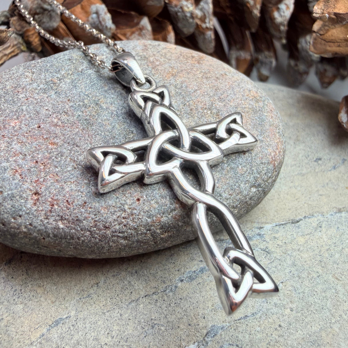A silver Celtic knot cross pendant on a matching chain, displayed on a smooth gray stone surrounded by pinecones.