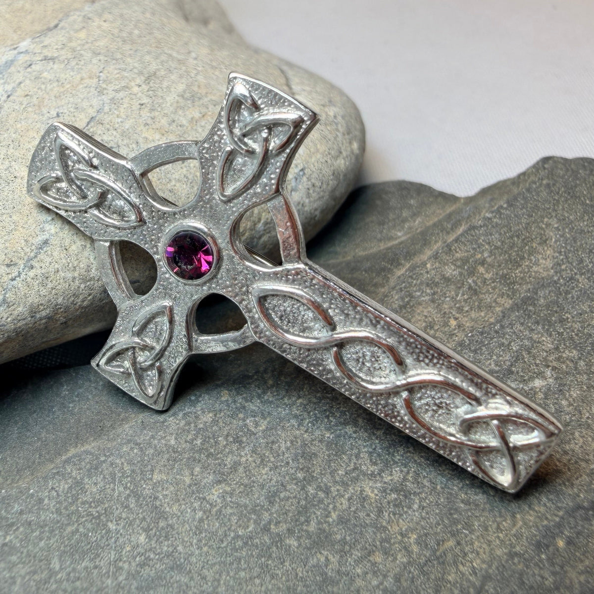 A silver Celtic cross brooch with a small pink gemstone in the center, resting on dark gray stones.