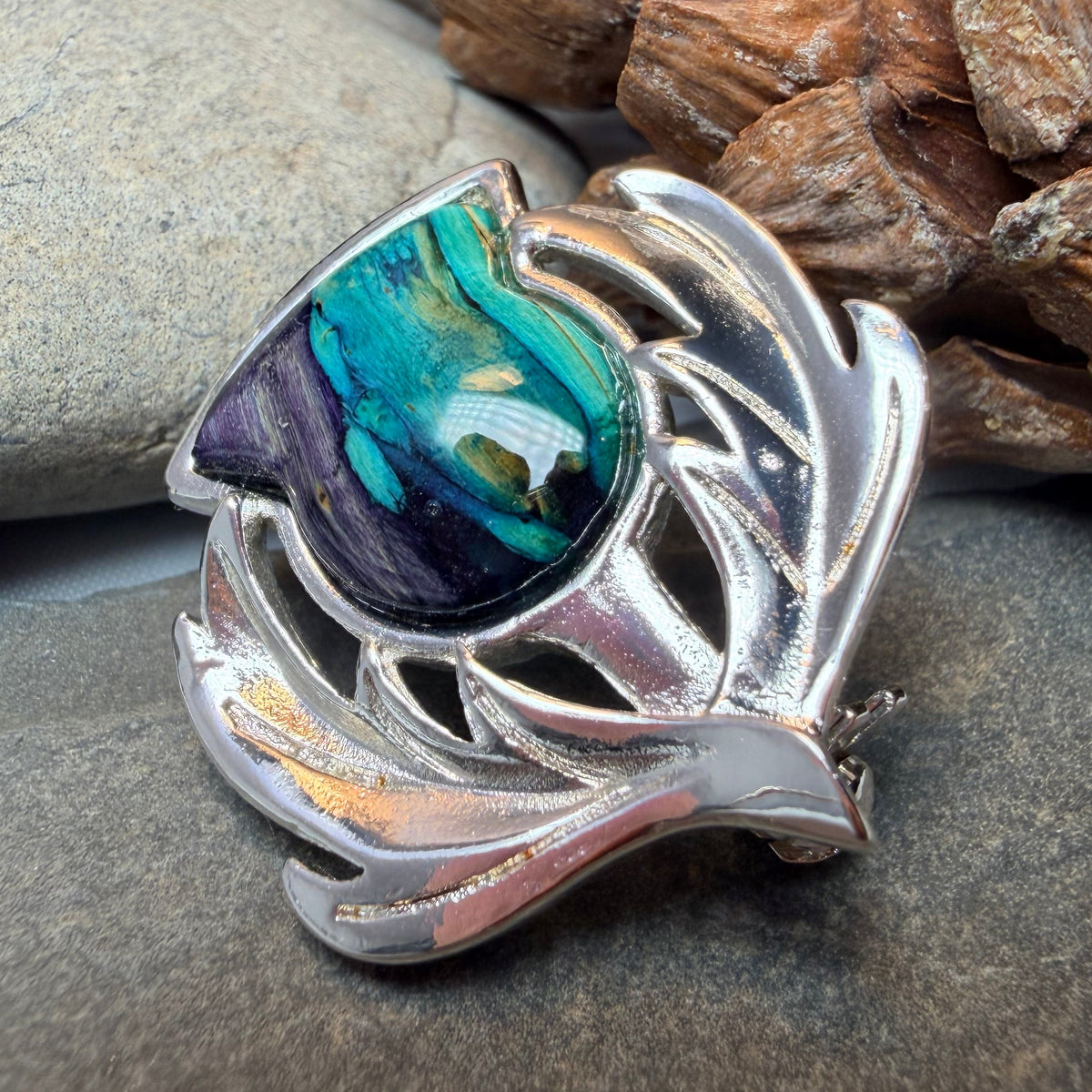 A silver-toned metal thistle brooch with a shield-shaped inlay featuring iridescent teal, blue, and purple heathergems displayed on a dark stone with a pinecone in the background.
