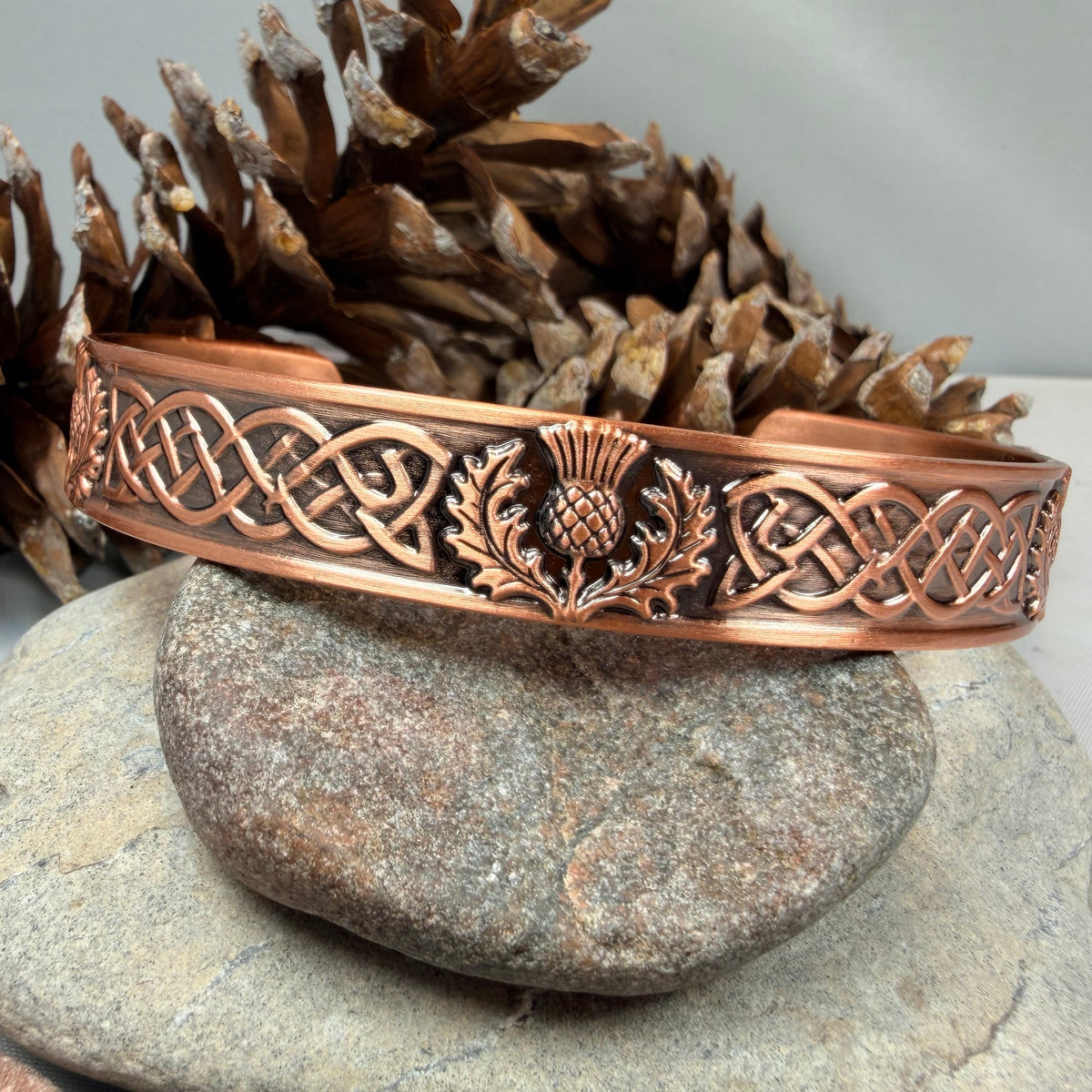 A copper-colored metal cuff bracelet with Celtic knotwork and a thistle motif, displayed on stacked stones with pine cones in the background.
