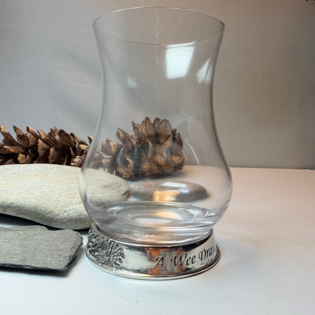 A clear glass whiskey tasting glass with a flared top and a silver-toned decorative base, photographed on a tabletop with stones, pinecones, and a gray cloth nearby.