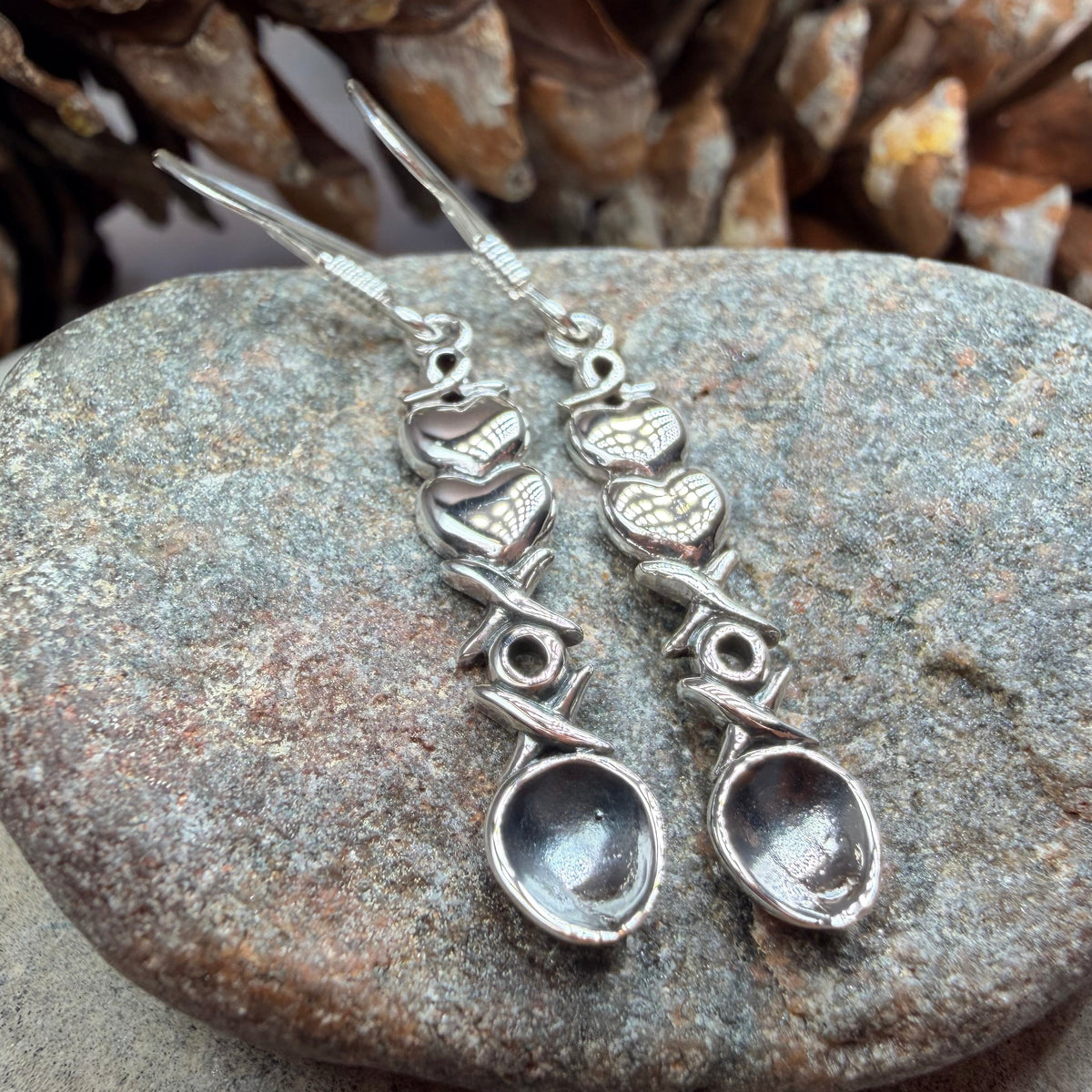 A pair of silver dangle earrings with small heart and swirl charms, each ending in a clear oval stone, displayed on a smooth gray stone with pinecones in the background.