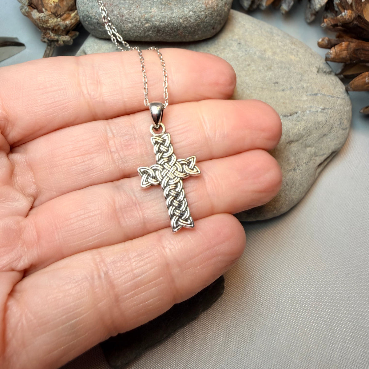 A sterling silver Celtic cross pendant on a delicate silver chain, held in a person's hand against a background of gray stones and pinecones.