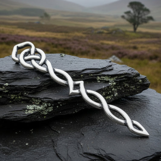 A silver-toned Celtic knot kilt pin with a twisted chain design, displayed on stacked natural stones with pinecones in the background.