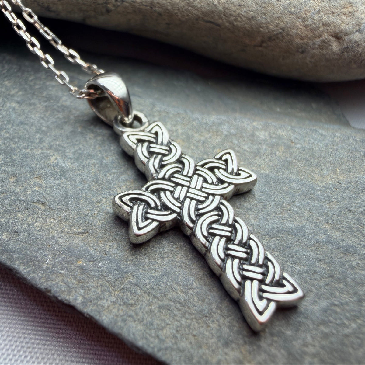 A silver Celtic knot cross pendant necklace resting on a textured gray stone surface with a smooth stone in the background.