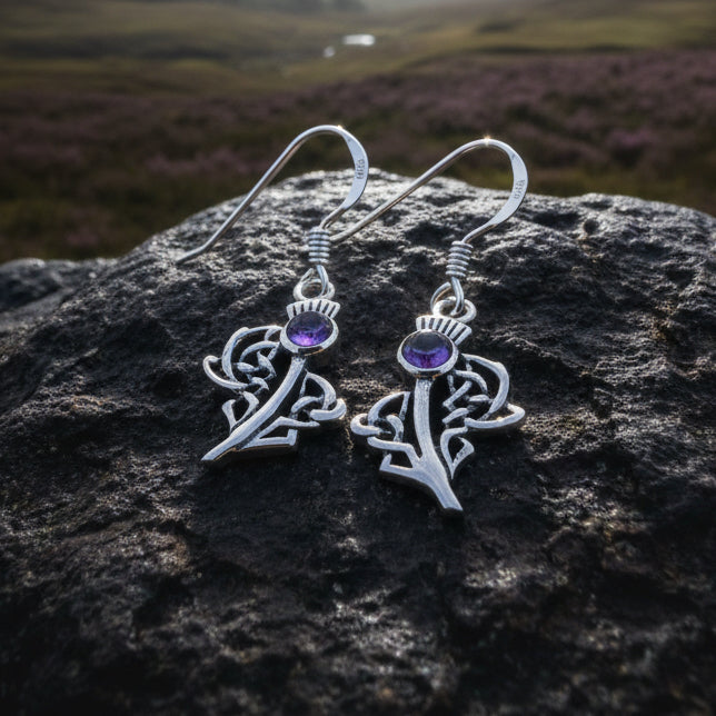 A pair of silver Celtic earrings with purple thistle accents, displayed on a smooth gray stone with pinecones in the background.