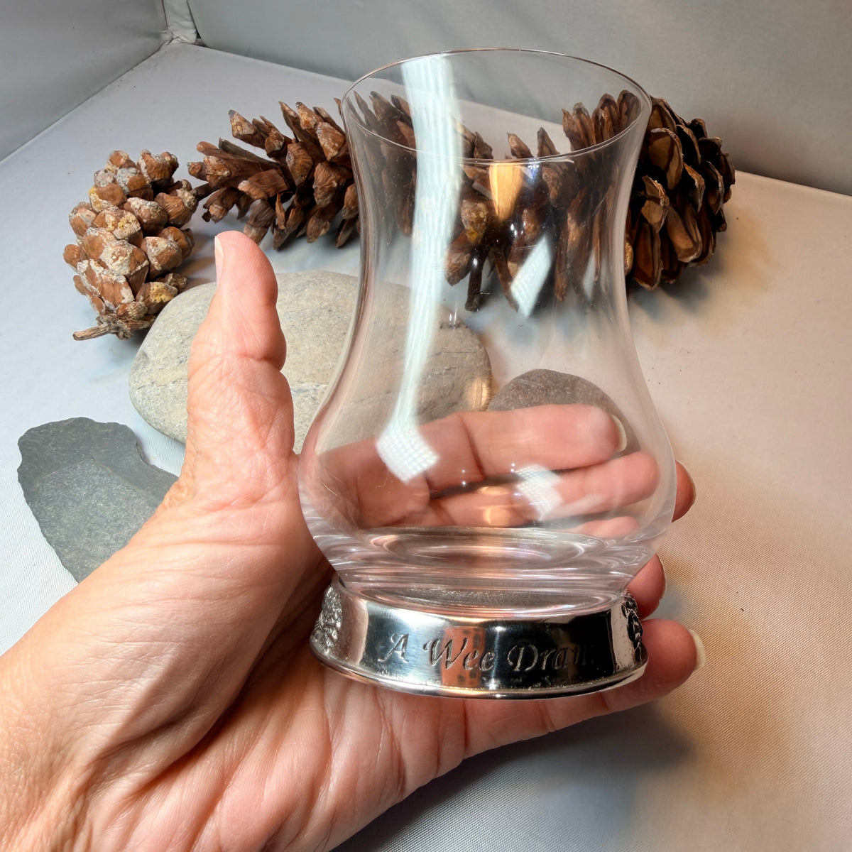 A hand holding a clear glass whisky glass with a silver metal base, with pinecones and stones 