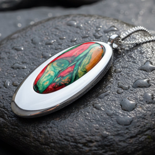 An oval Scottish Heathergems pendant necklace with a silver-toned metal frame and chain, featuring a colorful inlay of red, green, and white marbled or feather-like pattern, displayed on gray stones with pinecones.