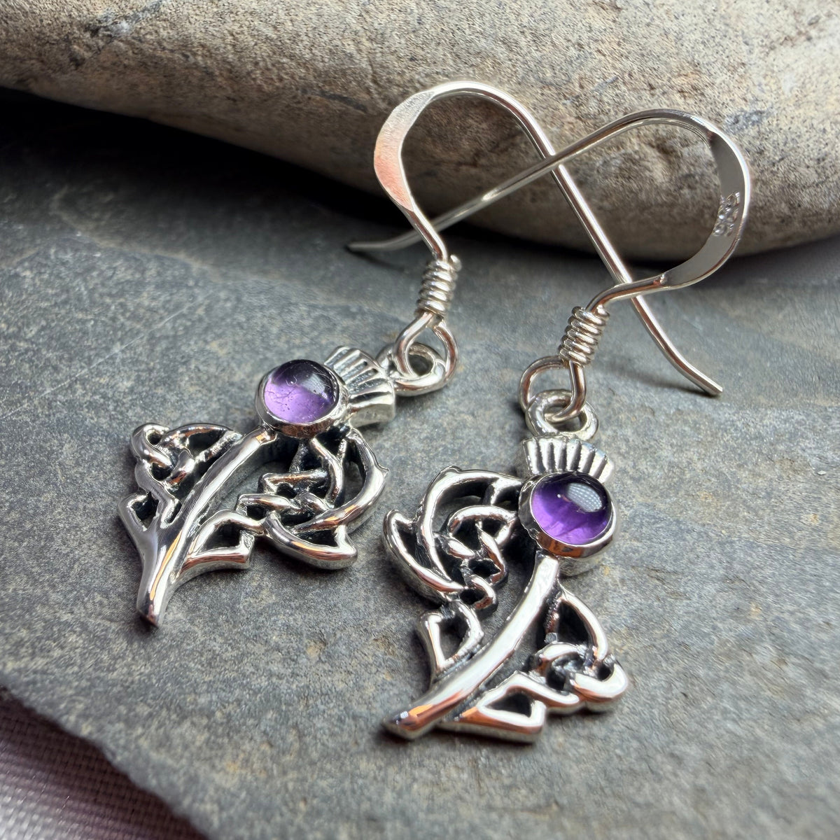 A pair of silver-toned dangle earrings with intricate Celtic knot thistle charms and small round purple gemstones, displayed on dark gray stones.
