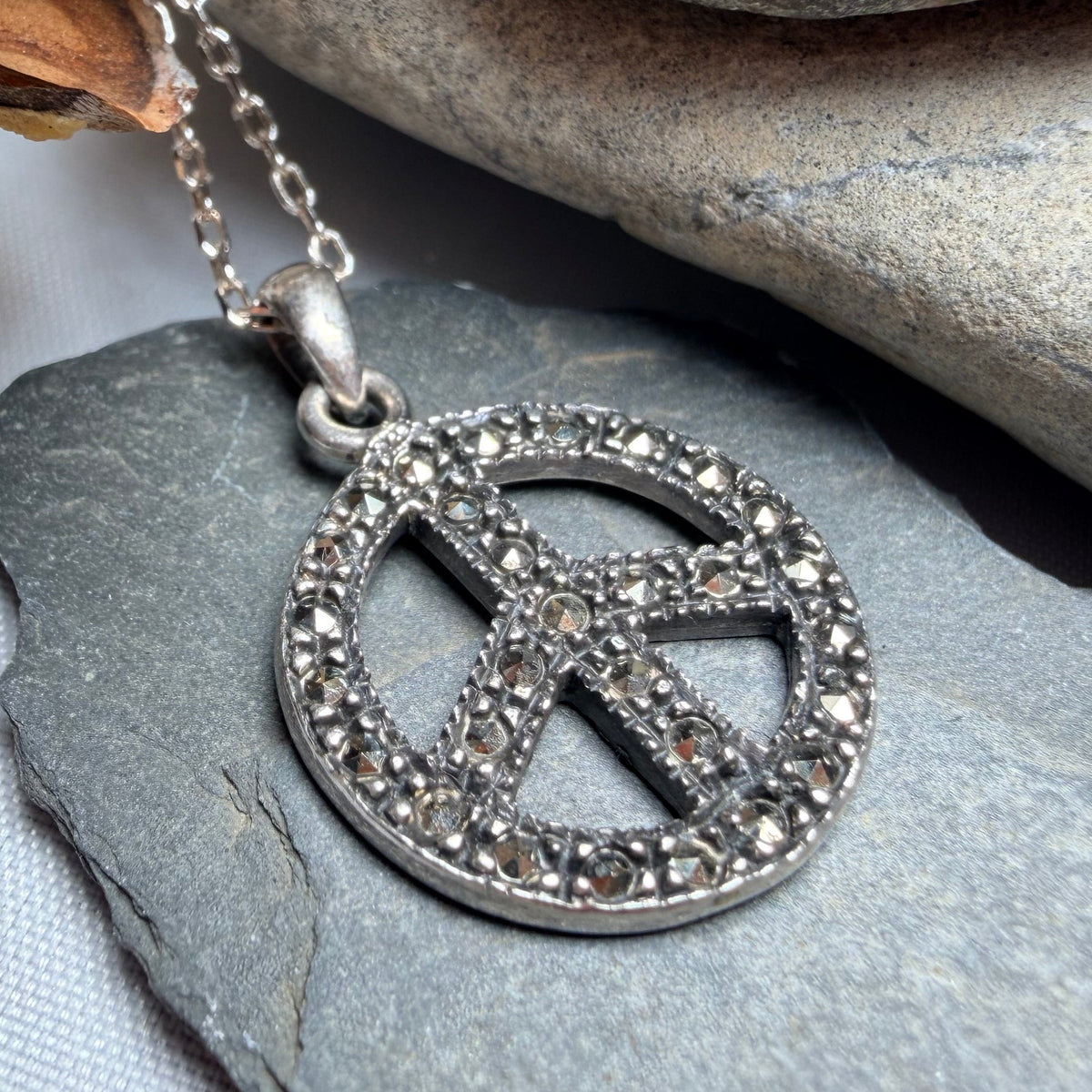A silver peace sign pendant necklace with sparkling clear stones, resting on a slate surface next to a pine cone and smooth rocks.