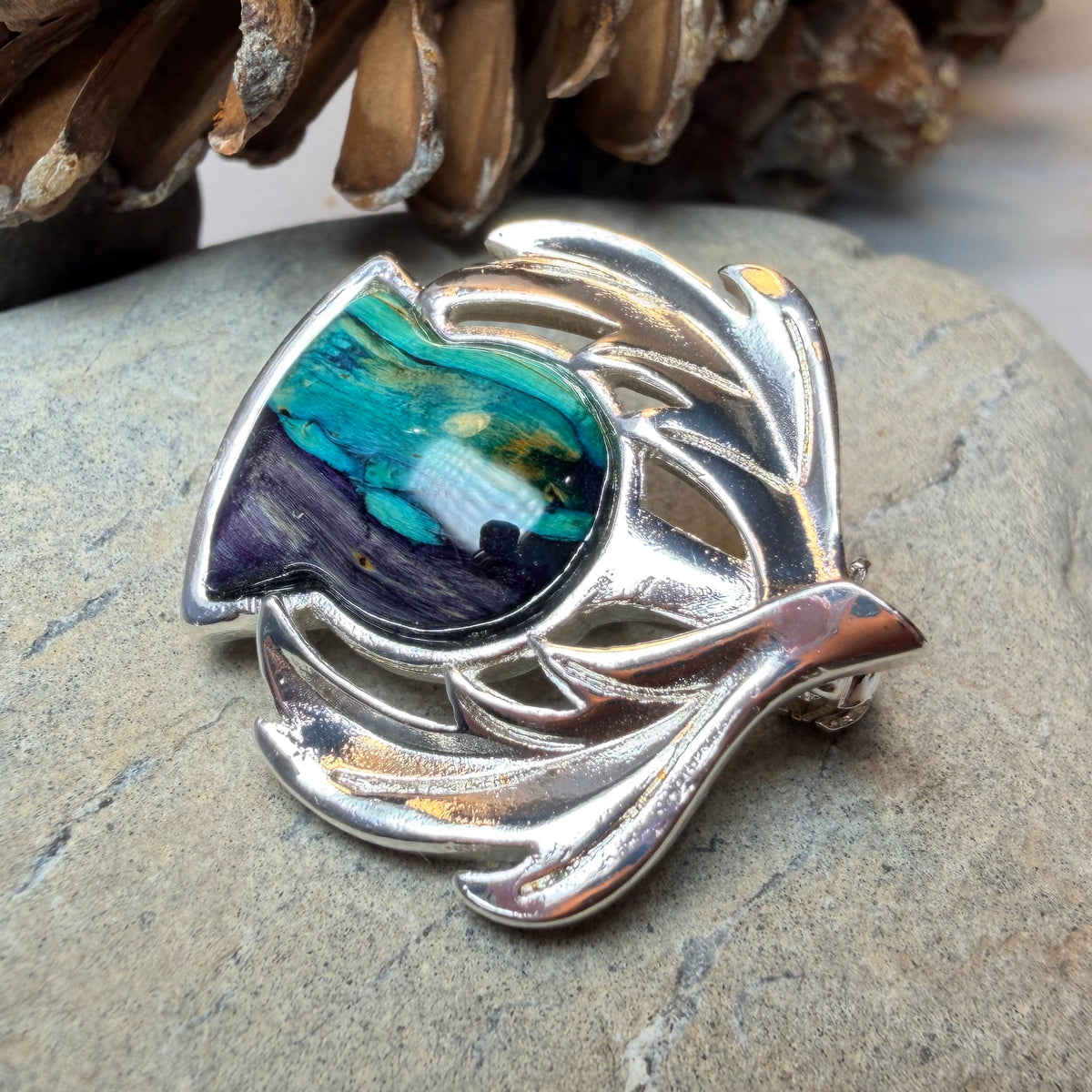 A silver-toned leaf-shaped brooch with a colorful inlay of turquoise, green, purple, and cream, resting on a gray stone with pinecones in the background made by Heathergems, Scotland.