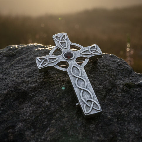 A silver Celtic cross brooch with an oxidized finish and a small dark red gemstone in the center, resting on a gray stone with pinecones in the background.
