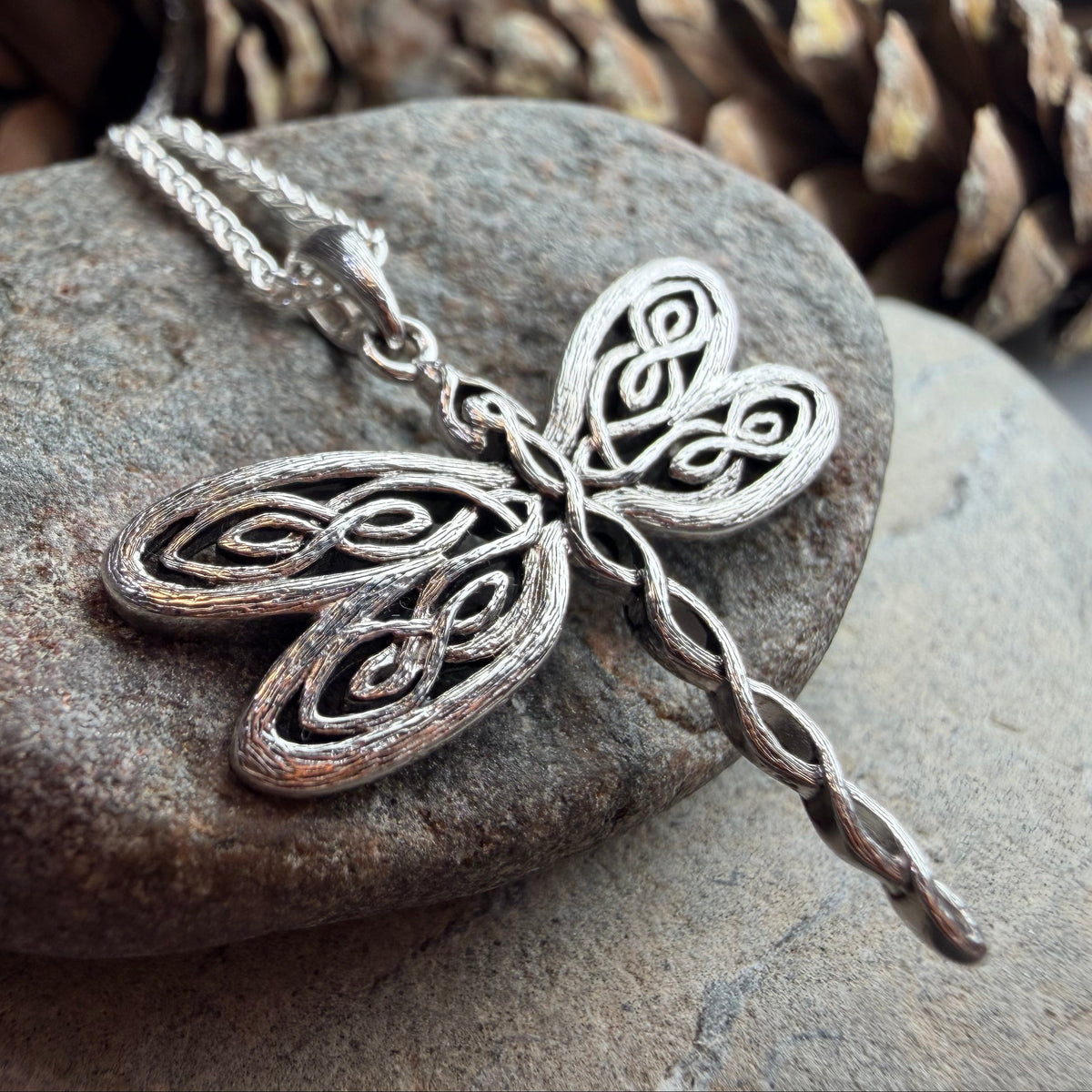 A silver-toned dragonfly pendant necklace resting on a smooth gray stone, with pinecones blurred in the background.