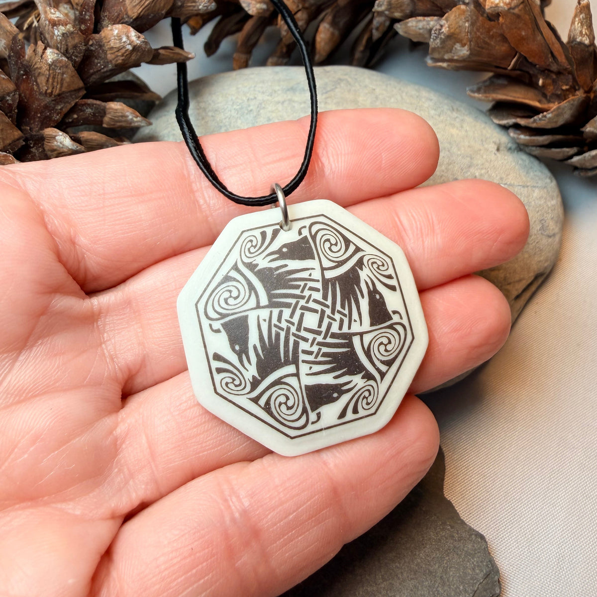 A hand holding an octagonal pendant necklace with a black cord and a white pendant featuring a black engraved knot-like design. The background includes pinecones and a stone.