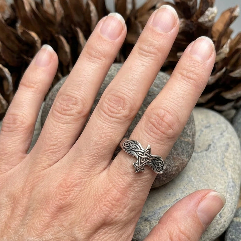 A silver Celtic knot ring worn on a finger, shown in a close-up shot with pine cones and a stone in the background.