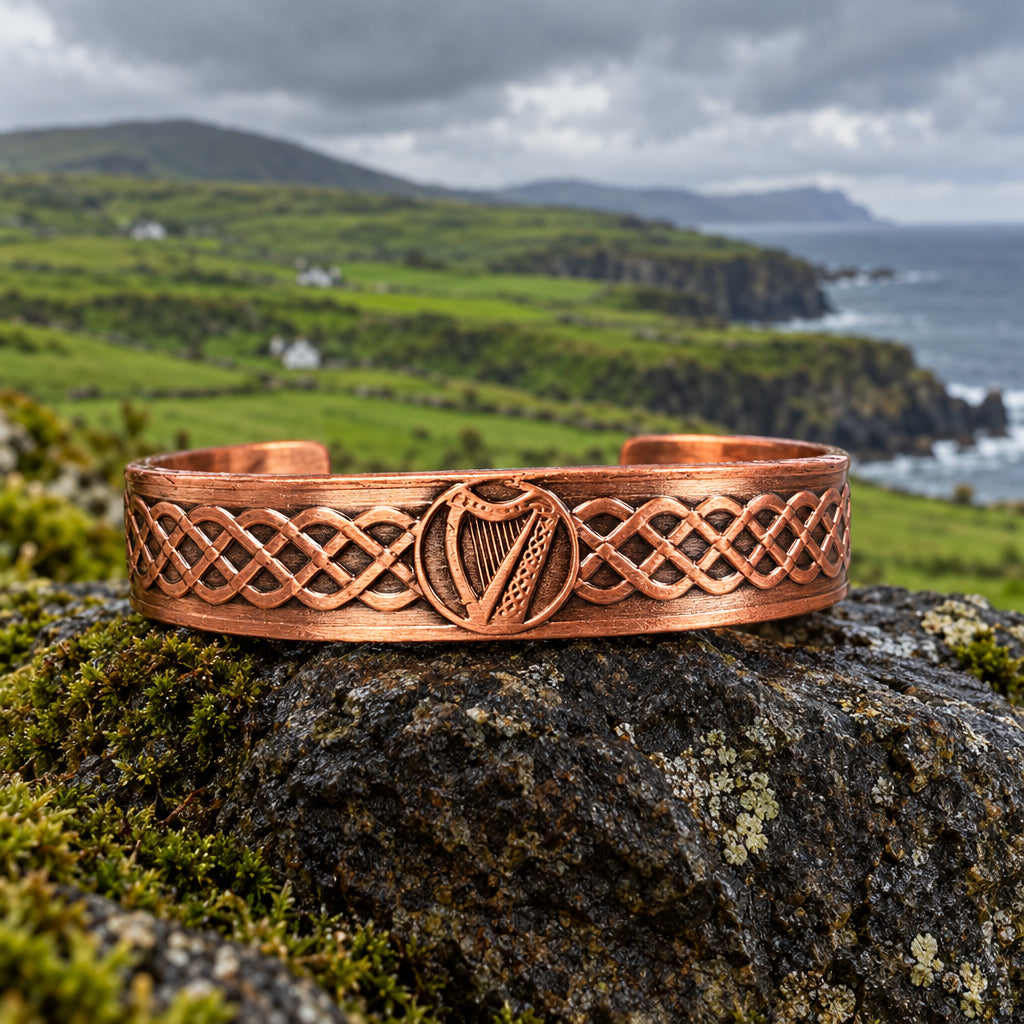 A copper-colored metal headband with Celtic knotwork and a harp motif, displayed on a stone with pinecones in the background.