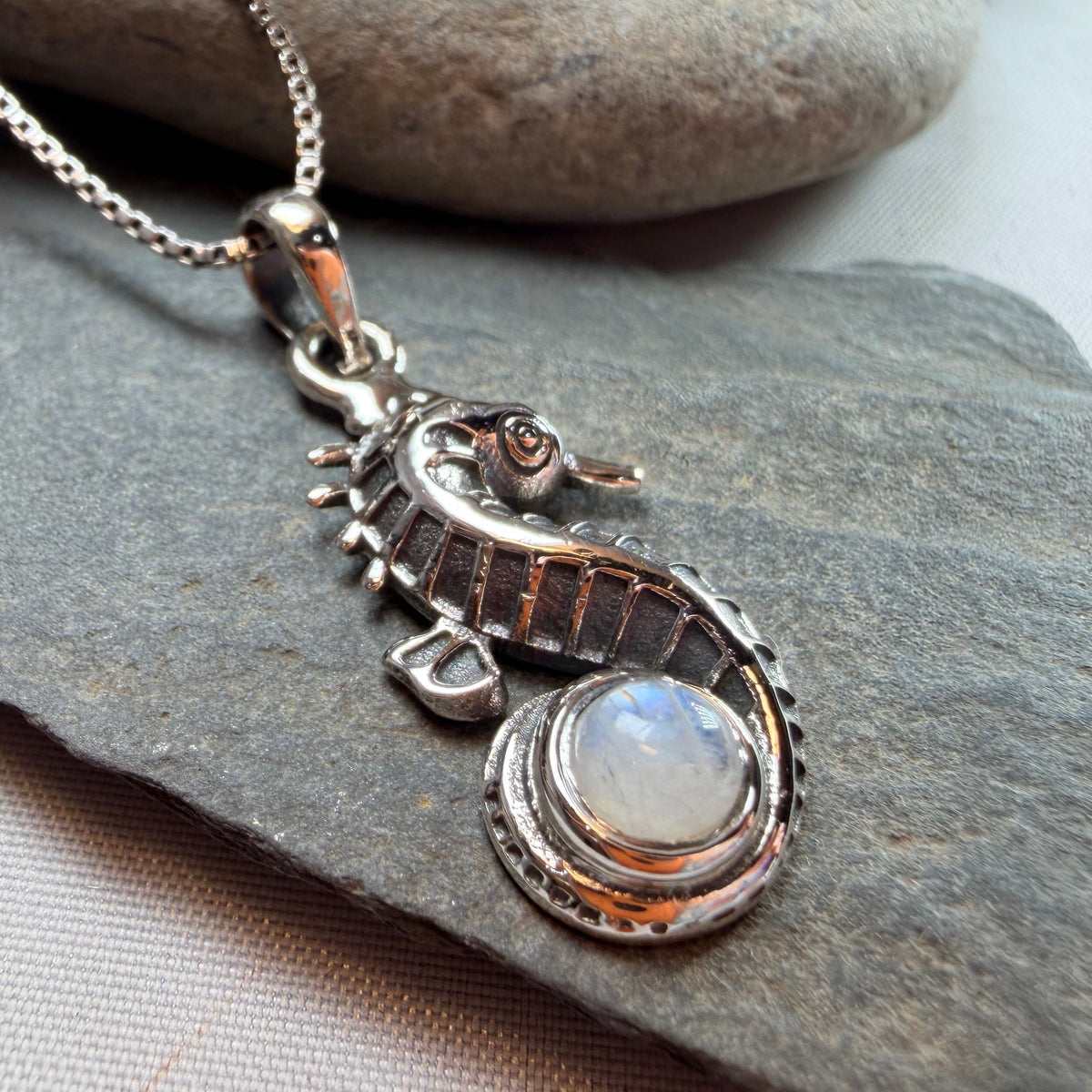 A silver-toned seahorse pendant necklace with a round iridescent moonstone, displayed on a gray slate and white fabric background.