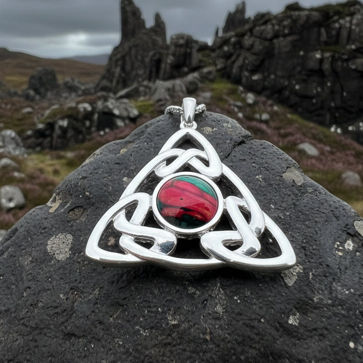 A silver Heathergems Celtic knot triquetra pendant necklace with a central oval inset featuring a red and green pattern, displayed on gray stones with pinecones. Made in Scotland