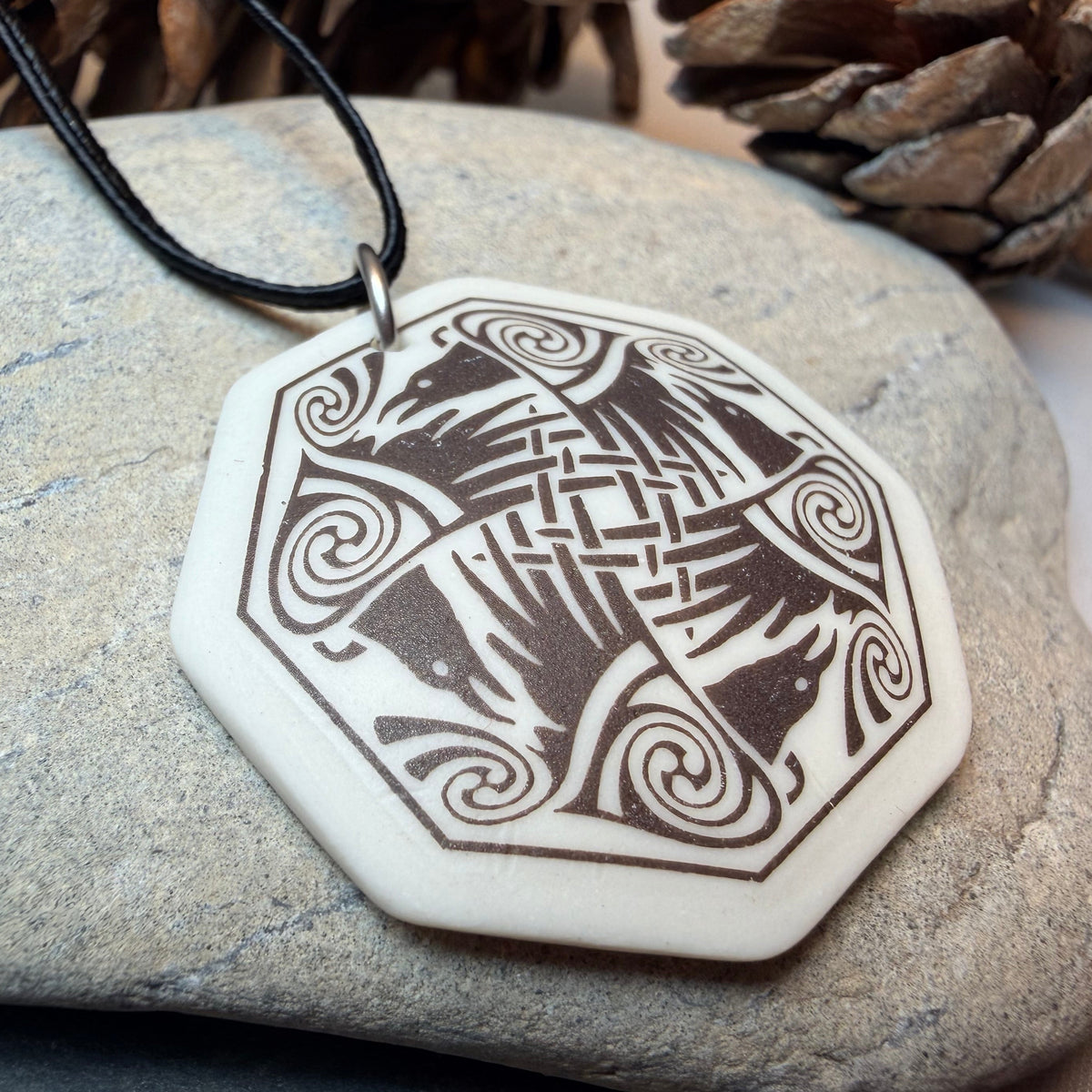 A hexagonal pendant necklace with a carved Norse raven design, resting on a smooth gray stone with pinecones in the background.
