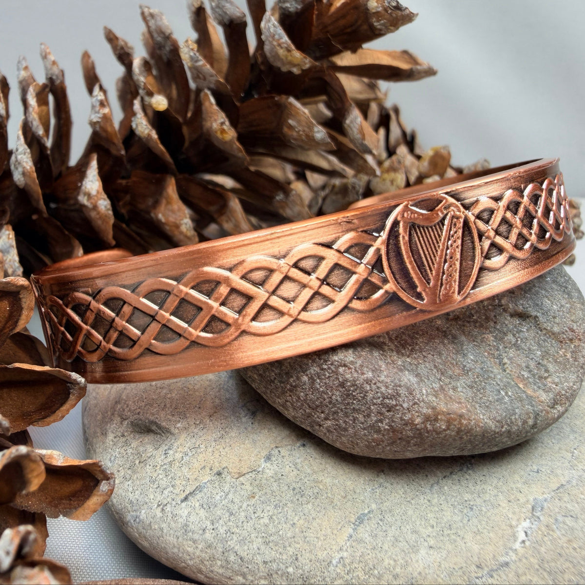 A copper-colored Celtic-style bangle bracelet with a harp and knotwork design, displayed on a stone with pinecones.