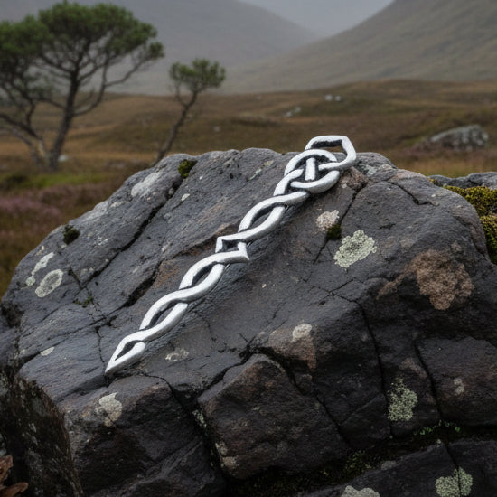 A silver Celtic knot kilt pin with a twisted chain design, displayed on stacked stones with pinecones in the background.